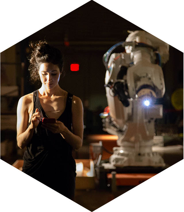 A woman facing forward looking at a phone with a large industrial robot behind her